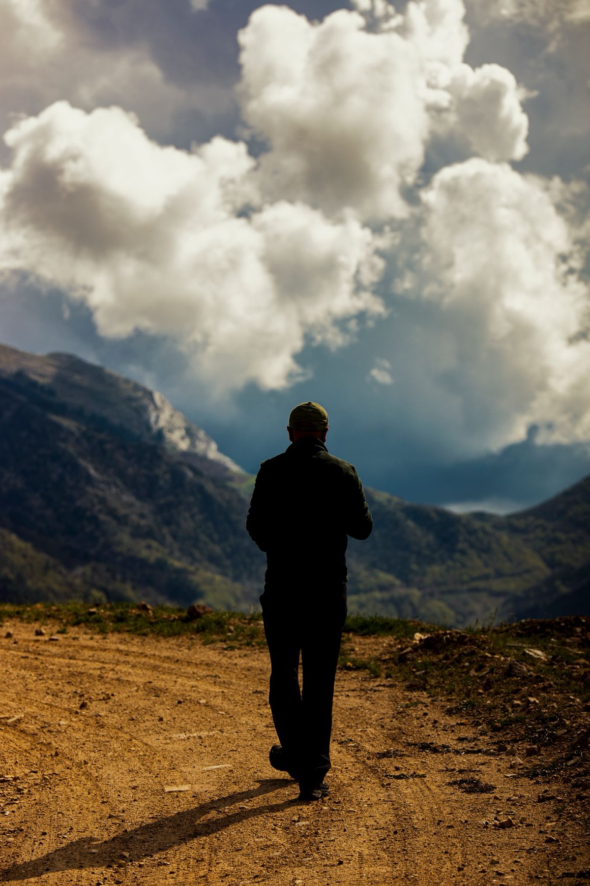 A lone figure on a dirt road looks out towards mountains under heavy cloud.